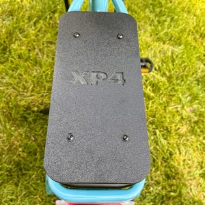 May include: A close-up view of the rear of a light blue bicycle. A black textured plate is mounted on the frame, with the letters "XP4" embossed on it. The bicycle has a red reflector and black pedals.