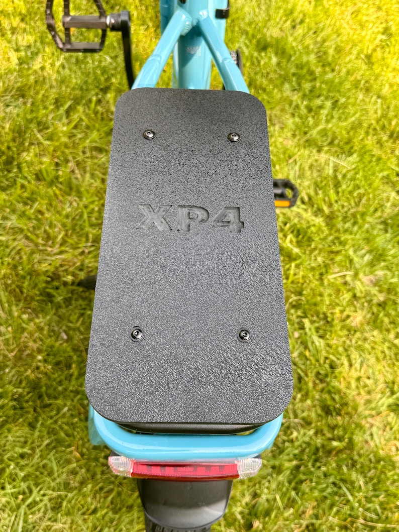 May include: A close-up view of the rear of a light blue bicycle. A black textured plate is mounted on the frame, with the letters "XP4" embossed on it. The bicycle has a red reflector and black pedals.