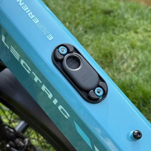May include: Close-up of a light blue electric bicycle frame with the word "LECTRIC" and "EXPERIENCE" printed on it. A black component with a silver button is attached to the frame with blue bolts. The words "galaxy smarttag" are printed on the black component.