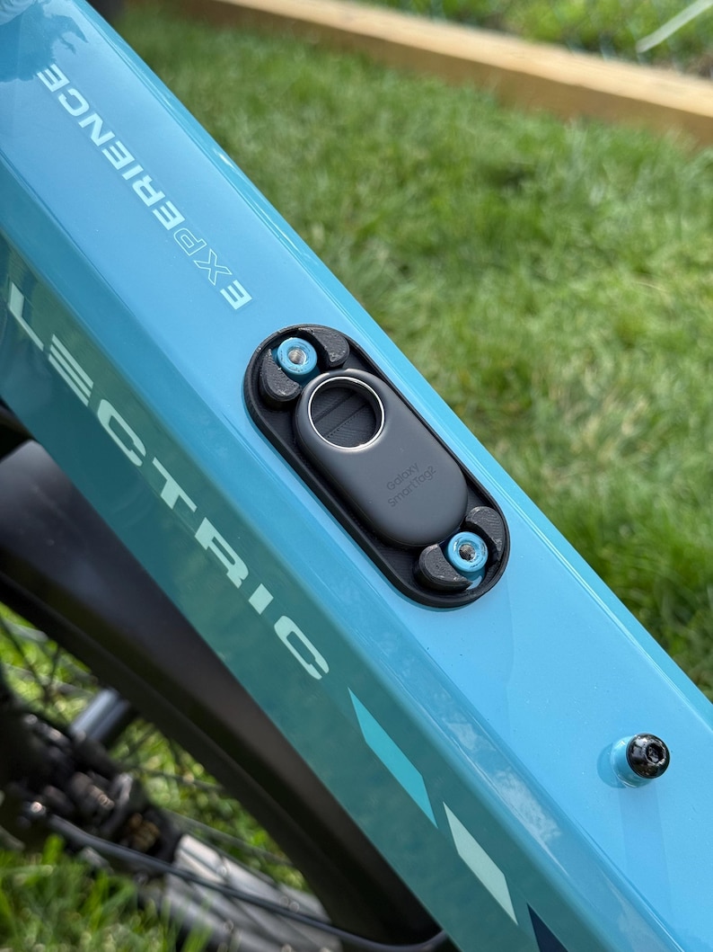 May include: Close-up of a light blue electric bicycle frame with the word "LECTRIC" and "EXPERIENCE" printed on it. A black component with a silver button is attached to the frame with blue bolts. The words "galaxy smarttag" are printed on the black component.