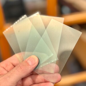 May include: A hand holding a fan of translucent, square-shaped plastic sheets. The sheets are slightly tinted and appear to be thin. The edges of the sheets are visible, and the overall effect is of a layered, semi-transparent material.