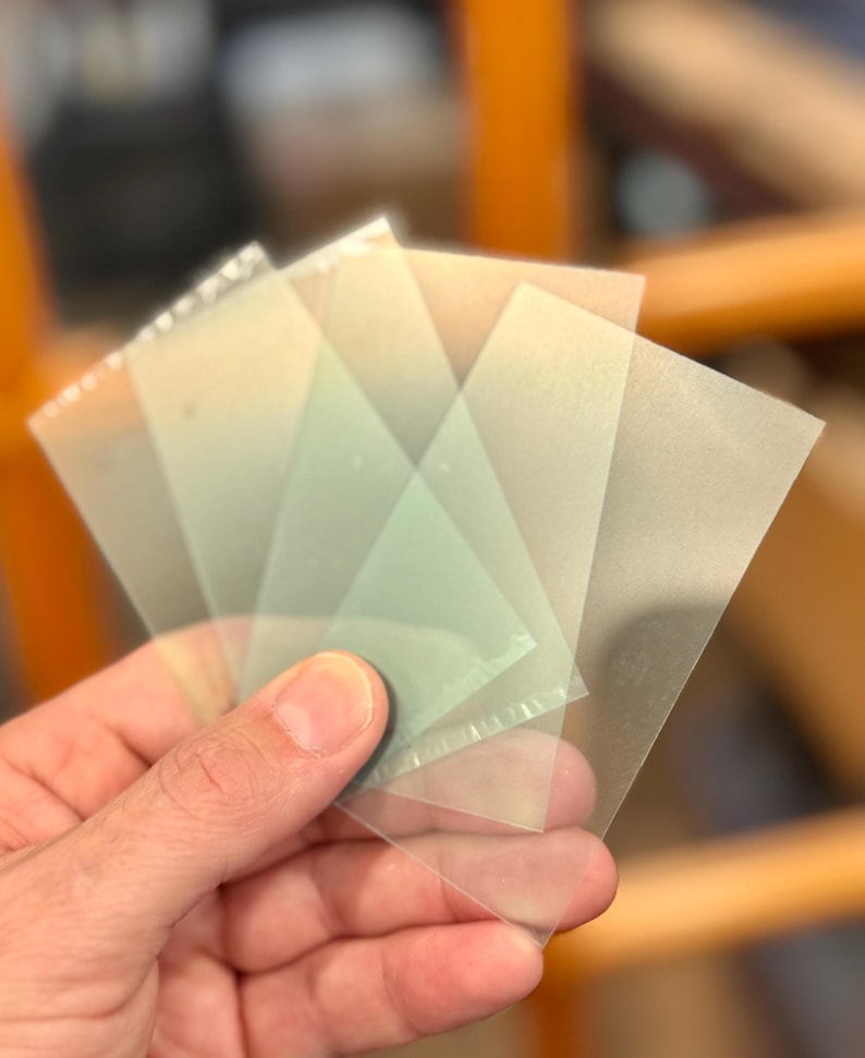 May include: A hand holding a fan of translucent, square-shaped plastic sheets. The sheets are slightly tinted and appear to be thin. The edges of the sheets are visible, and the overall effect is of a layered, semi-transparent material.