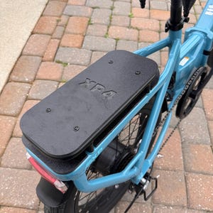 May include: A light blue electric bicycle with a black seat and rear rack. The rear tire has a black fender and a red reflector. The black rack has the text "XP4" on it.