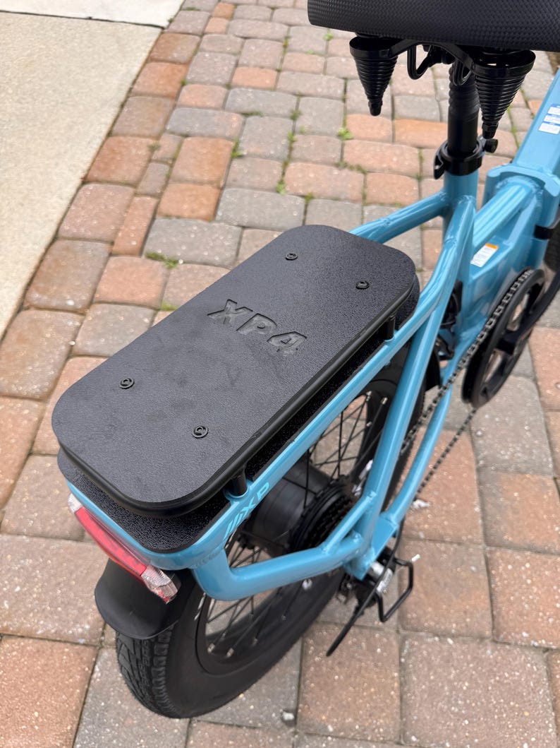 May include: A light blue electric bicycle with a black seat and rear rack. The rear tire has a black fender and a red reflector. The black rack has the text "XP4" on it.