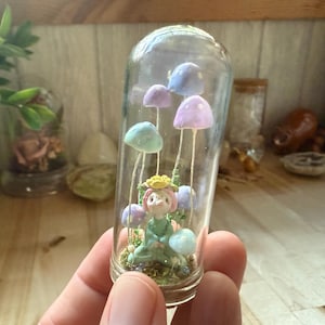 Puede incluir: Una pequeña figura de hada sentada entre setas de colores pastel en una cúpula de vidrio. El hada lleva un vestido verde y una corona de flores amarillas.