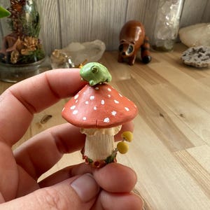 May include: A whimsical miniature mushroom figurine with a red cap and white spots, topped with a small green frog. The mushroom has a cream-colored stem and small yellow mushrooms at the base. A charming, handcrafted decorative item.