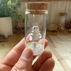 Puede incluir: Una pequeña figurita blanca de una criatura está dentro de un frasco de vidrio transparente con un tapón de corcho. El frasco está lleno de purpurina iridiscente.