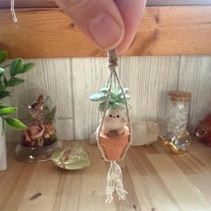 May include: A small, white ceramic animal figurine with a green plant growing from its head, hanging from a macrame plant hanger. The figurine is in a small terracotta pot.