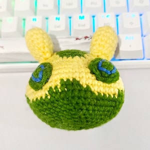 May include: A crocheted plush toy head in yellow and green, with blue detailing. The handmade item has prominent ears and facial features. The toy's design is clearly visible, highlighting the craftsmanship of the item.