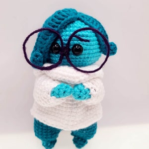May include: A blue crocheted character with purple glasses and a white crocheted sweater. The character has black eyes and a sad expression.