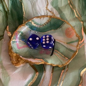 May include: A decorative, hand-painted dish with a gold-painted rim, holding two blue dice with white dots. The dish has a green and gold design, set against a green and white patterned background. A unique piece for home decor.