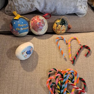 May include: A collection of Christmas ornaments, including a round ornament with a blue background and the text "Merry Christmas", a round ornament with a red and white background, a round ornament with a white background and a snowman design, and a white egg-shaped ornament with a blue bunny design. There are also several colorful pipe cleaner candy canes.