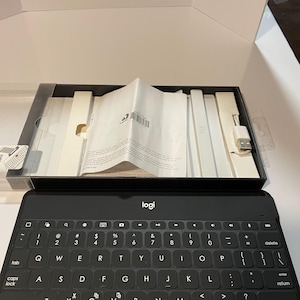 May include: A black Logi keyboard with white lettering and symbols is displayed in an open box. The keyboard is in front of a clear plastic case containing a USB cable and paperwork. The box is white.