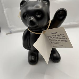 May include: A matte black ceramic bear figurine with one arm raised in a waving gesture. A tan string is tied around its neck, holding a white paper tag that reads: Boogie: Boogie, Dancer, Night Fever, Shaker, Two-Step. Birthdate: 9-6-02.