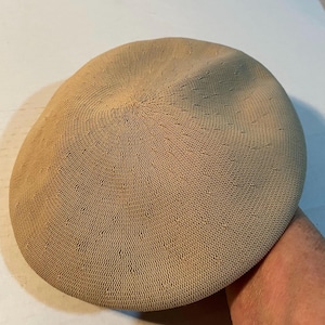 May include: A beige beret crafted from a fine, tightly woven knit fabric. The hat features a circular, flat-topped design with a subtle, uniform texture across the entire surface, showcasing a consistent tan color and a structured, rounded shape.
