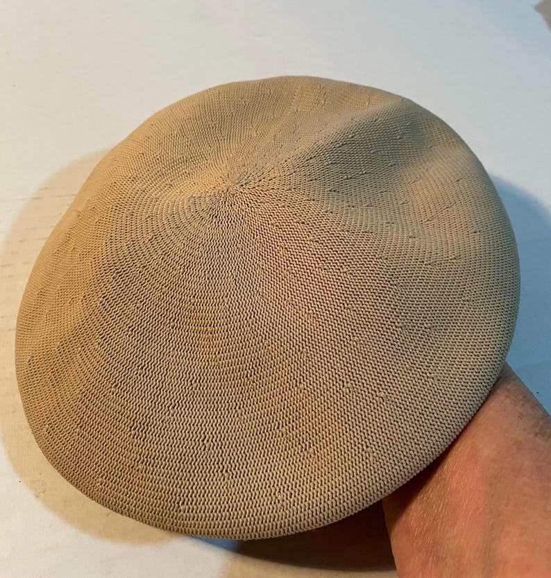 May include: A beige beret crafted from a fine, tightly woven knit fabric. The hat features a circular, flat-topped design with a subtle, uniform texture across the entire surface, showcasing a consistent tan color and a structured, rounded shape.