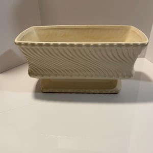 May include: Cream-coloured rectangular planter with a pedestal base. The planter has a textured, swirling pattern on the sides and a beaded edge along the top. The base is a solid, rectangular shape.
