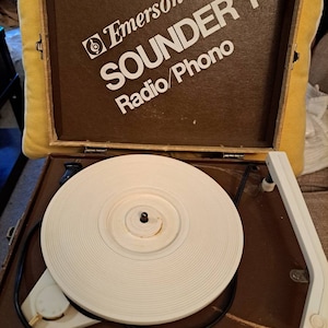 Pu&ograve; includere: Un giradischi vintage Emerson Sounder 1 Radio/Phono di colore marrone con un piatto girevole bianco e un cavo nero. Il giradischi &egrave; aperto e il piatto girevole &egrave; visibile.