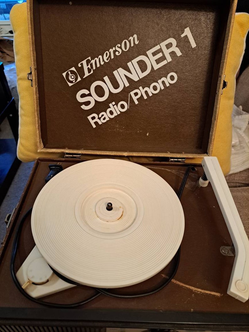 Pu&ograve; includere: Un giradischi vintage Emerson Sounder 1 Radio/Phono di colore marrone con un piatto girevole bianco e un cavo nero. Il giradischi &egrave; aperto e il piatto girevole &egrave; visibile.