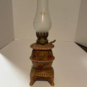 May include: A decorative oil lamp with a frosted glass globe and a brown base shaped like a stove. The base features red rose accents and a brass-colored handle and details. The lamp sits on a small, rectangular base.