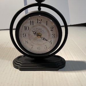 May include: A black metal table clock featuring a circular frame topped with a small bird figurine. The clock face has black Arabic numerals, ornate hands, and the text PARIS, EST 1910, 18, RUE PERREULE printed in the center on a cream background.