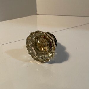 May include: A clear, faceted glass doorknob with a brass-coloured centre and a dark, possibly black, base. The knob has a starburst design in the centre and is resting on a white surface.