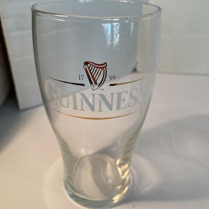 May include: A clear Guinness pint glass with the Guinness logo in gold and the harp symbol in red and gold. The glass has a wide top and tapers down to a smaller base. The text "17 59" is above the logo.