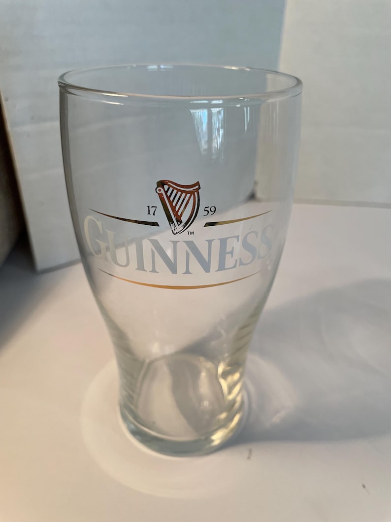 May include: A clear Guinness pint glass with the Guinness logo in gold and the harp symbol in red and gold. The glass has a wide top and tapers down to a smaller base. The text "17 59" is above the logo.