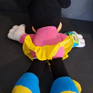 May include: A plush Minnie Mouse toy wearing a pink dress with a yellow skirt and a yellow bow with white polka dots. She has blue and yellow shoes and a green and blue watch on her wrist.