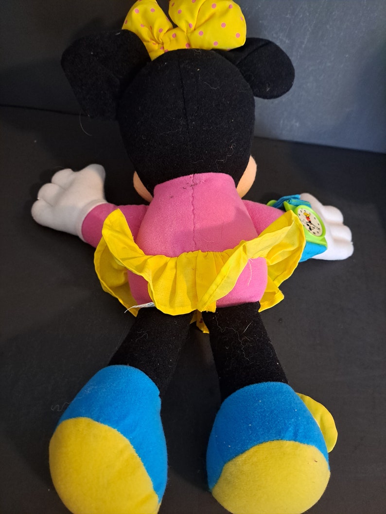 May include: A plush Minnie Mouse toy wearing a pink dress with a yellow skirt and a yellow bow with white polka dots. She has blue and yellow shoes and a green and blue watch on her wrist.