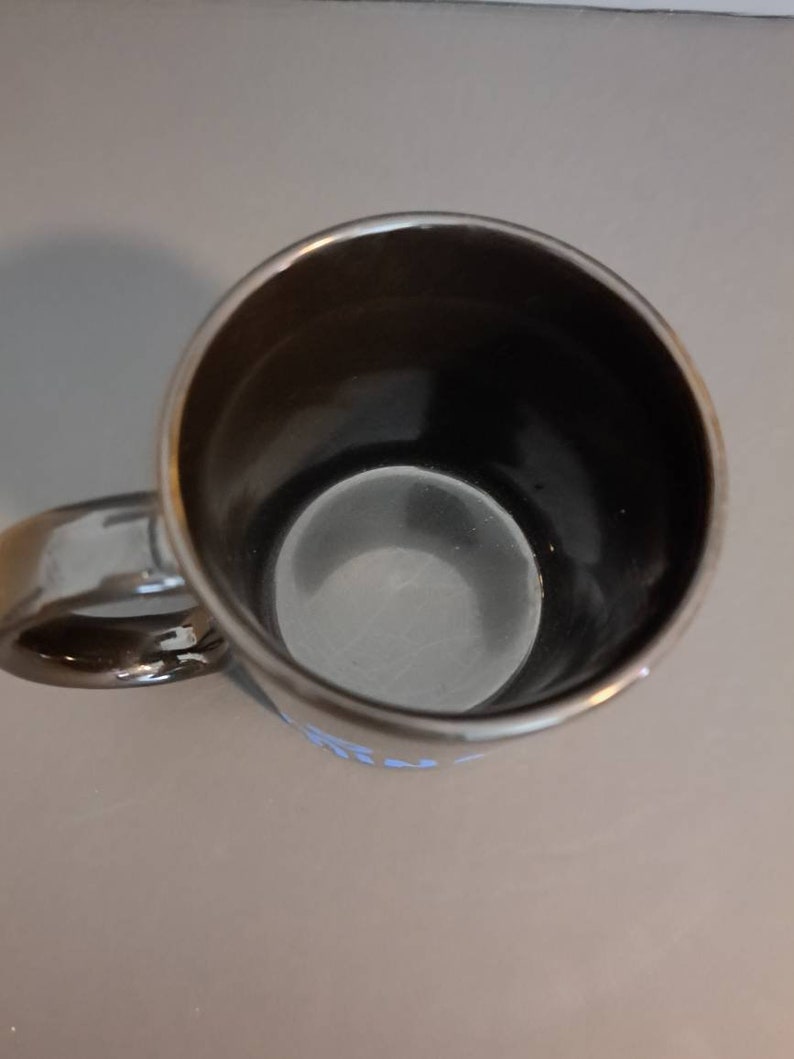Peut inclure: Une tasse en c&eacute;ramique noire avec un bord et une anse argent&eacute;s. L'int&eacute;rieur de la tasse est sombre et l'ext&eacute;rieur a une finition brillante. La tasse est vide et pos&eacute;e sur une surface gris clair.