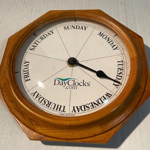 May include: An octagonal wall clock with a light brown wood frame and a white face divided into seven sections labeled with the days of the week. A single black hand points to Tuesday. The center features the text DayClocks.com and Patent #D433348.