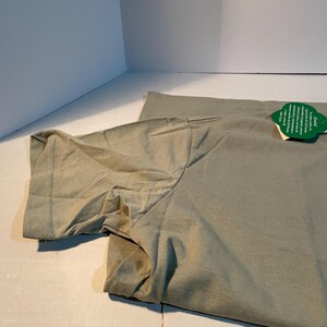 May include: A sage green short-sleeved t-shirt featuring a round neckline and a green leaf-shaped tag attached to the collar. The fabric has a visible weave and a smooth texture, with the sleeve folded slightly to show the hemmed edge.