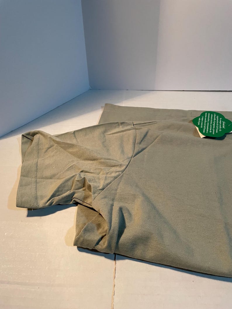 May include: A sage green short-sleeved t-shirt featuring a round neckline and a green leaf-shaped tag attached to the collar. The fabric has a visible weave and a smooth texture, with the sleeve folded slightly to show the hemmed edge.