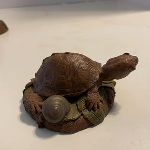 May include: A brown turtle figurine with a textured shell, positioned on a base of green leaves and a small gray snail. The base features the embossed text "W.W. 1993". The turtle is depicted with its head extended forward and legs resting on the foliage.