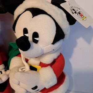 Puede incluir: Un Mickey Mouse de peluche vestido con un traje de Pap&aacute; Noel rojo y blanco, sosteniendo una bolsa de regalo verde. El juguete de peluche tiene una cara en blanco y negro con ojos negros y un borde de piel blanca alrededor del sombrero. La etiqueta adjunta al juguete de peluche dice "Mickey Mouse Co."