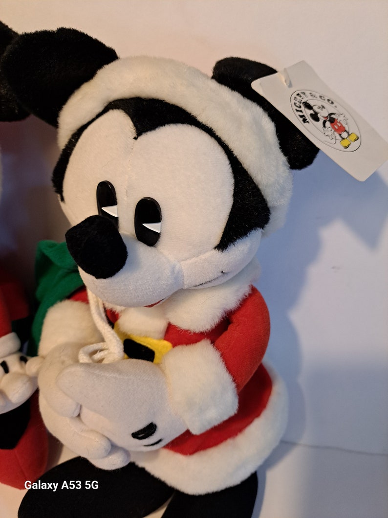 Puede incluir: Un Mickey Mouse de peluche vestido con un traje de Pap&aacute; Noel rojo y blanco, sosteniendo una bolsa de regalo verde. El juguete de peluche tiene una cara en blanco y negro con ojos negros y un borde de piel blanca alrededor del sombrero. La etiqueta adjunta al juguete de peluche dice "Mickey Mouse Co."