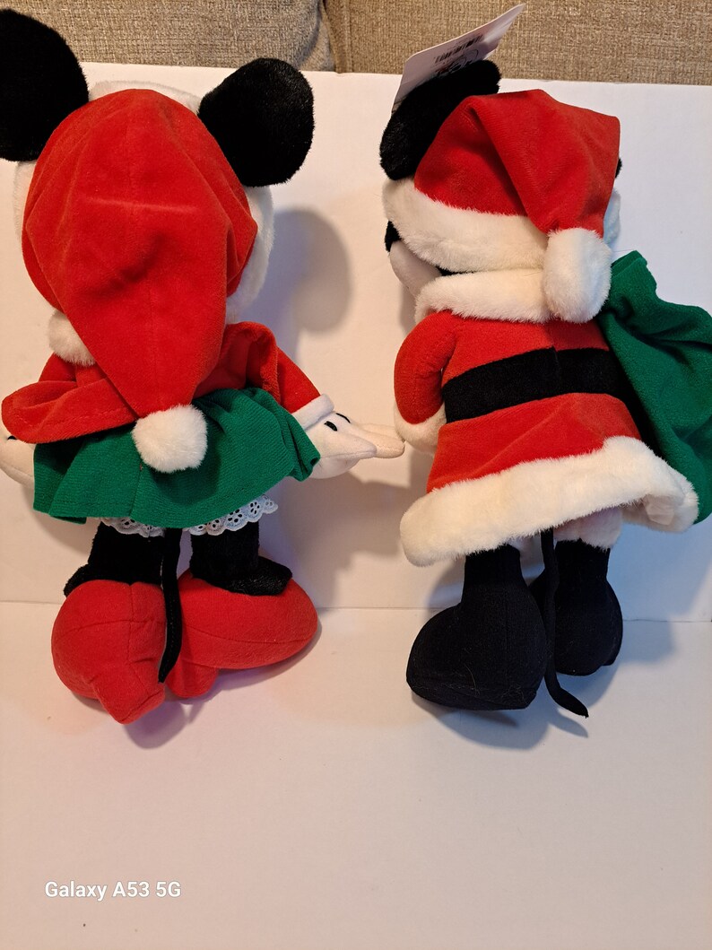 Puede incluir: Un par de juguetes de peluche de Mickey Mouse y Minnie Mouse con tem&aacute;tica navide&ntilde;a. Mickey est&aacute; vestido de Pap&aacute; Noel con un traje rojo, cintur&oacute;n negro y ribetes blancos. Minnie lleva un gorro de Pap&aacute; Noel rojo, un vestido verde y zapatos blancos.