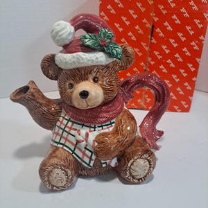 May include: A ceramic teapot shaped like a brown teddy bear wearing a red and white Santa hat, a red scarf, and a plaid shirt. The teapot has a red and white candy cane design on the front.
