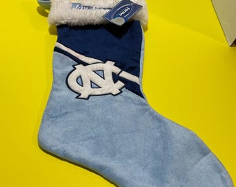 Può includere: Un calzino di Natale blu e bianco con il logo dei Tar Heels dell'Università della Carolina del Nord. Il calzino ha un polsino in pelliccia sintetica bianca e un design a righe blu e bianche. Il calzino ha un'etichetta "Forzieri" attaccata.