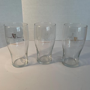 May include: Three clear pint glasses with the Guinness logo and harp emblem. The glasses have a curved shape and a gold accent line. The Guinness logo is printed in gold. The glasses are empty.