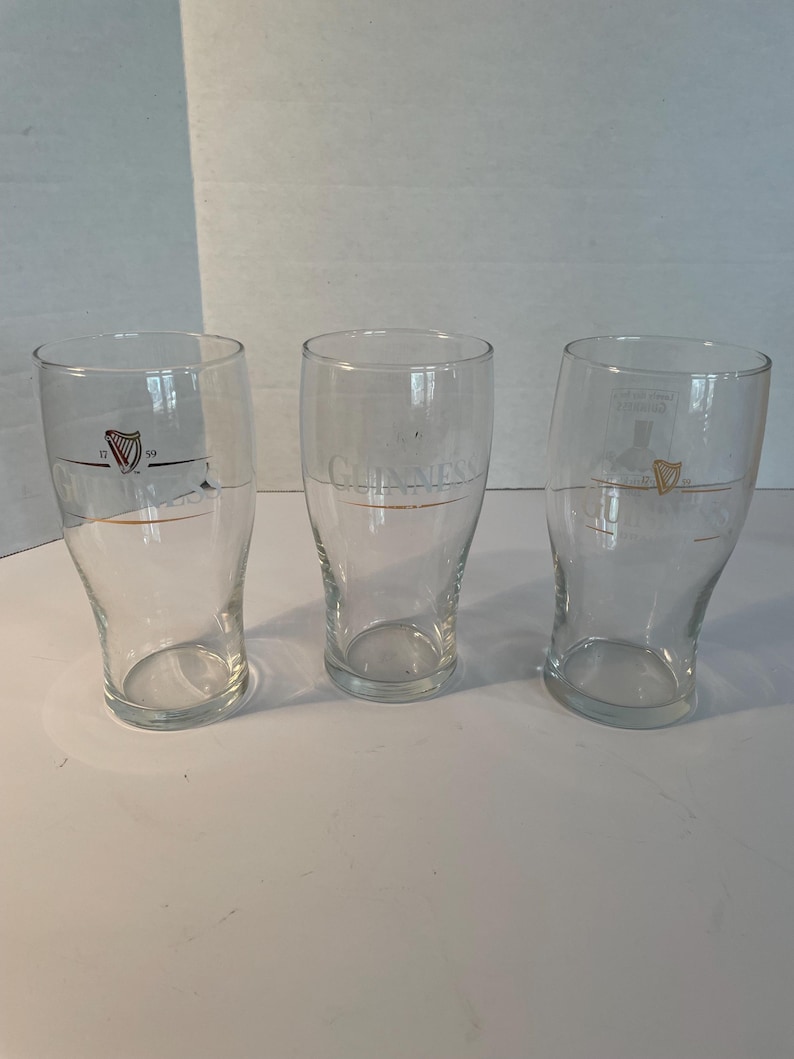 May include: Three clear pint glasses with the Guinness logo and harp emblem. The glasses have a curved shape and a gold accent line. The Guinness logo is printed in gold. The glasses are empty.