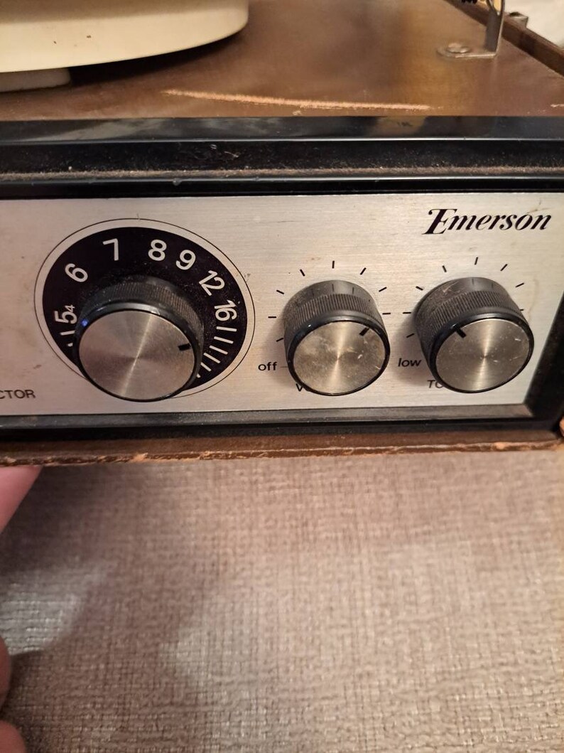 Pu&ograve; includere: Primo piano di un giradischi Emerson vintage con una piastra frontale argentata e tre manopole. Le manopole sono etichettate "off", "low" e "tone". Il giradischi ha una finitura in legno di colore marrone e nero.