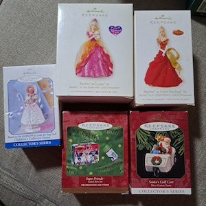 May include: Set of six Hallmark Keepsake Christmas ornaments. The ornaments feature Barbie as Liana in Barbie and the Diamond Castle, Barbie as Eden Starling in Barbie in a Christmas Carol, Barbie as Little Bo Peep, Super Friends Lunch Box Set, and Santa's Golf Cart. The ornaments are packaged in boxes with colorful illustrations and text.