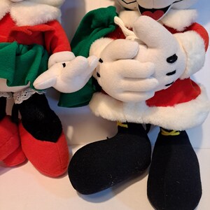 Puede incluir: Un par de personajes de peluche de Mickey Mouse con tem&aacute;tica navide&ntilde;a. Mickey lleva un traje de Pap&aacute; Noel rojo y blanco con zapatos negros. Minnie lleva un vestido rojo y blanco con una falda verde y zapatos rojos.
