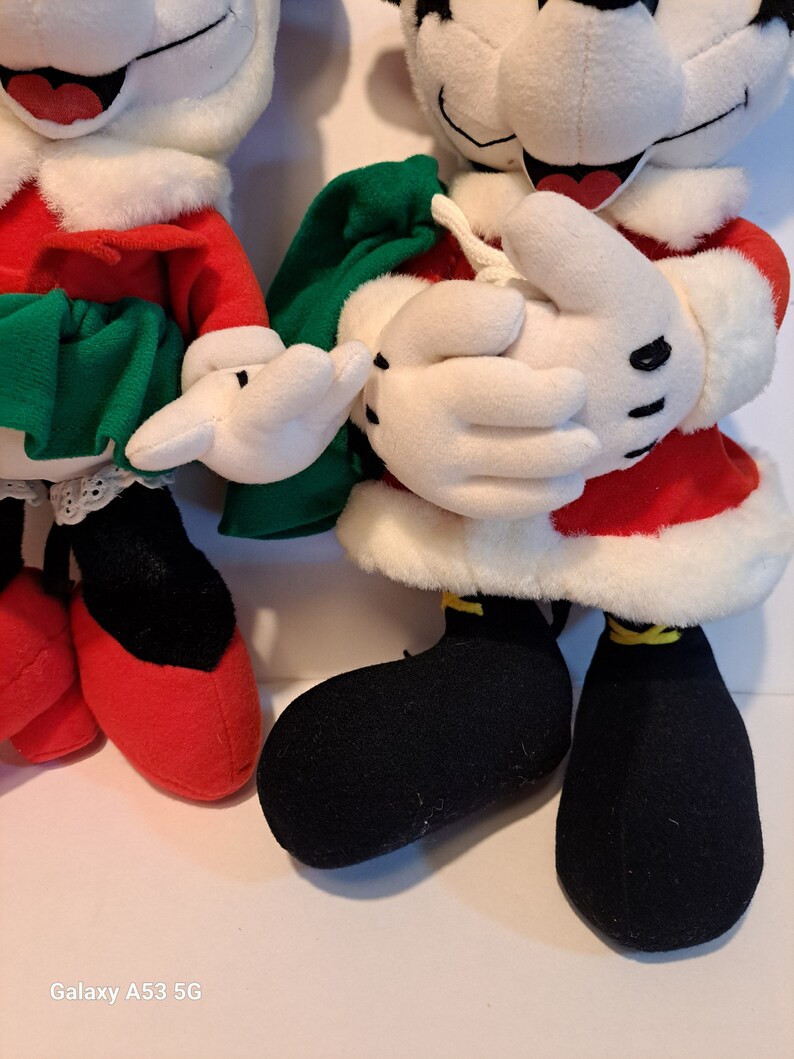 Puede incluir: Un par de personajes de peluche de Mickey Mouse con tem&aacute;tica navide&ntilde;a. Mickey lleva un traje de Pap&aacute; Noel rojo y blanco con zapatos negros. Minnie lleva un vestido rojo y blanco con una falda verde y zapatos rojos.