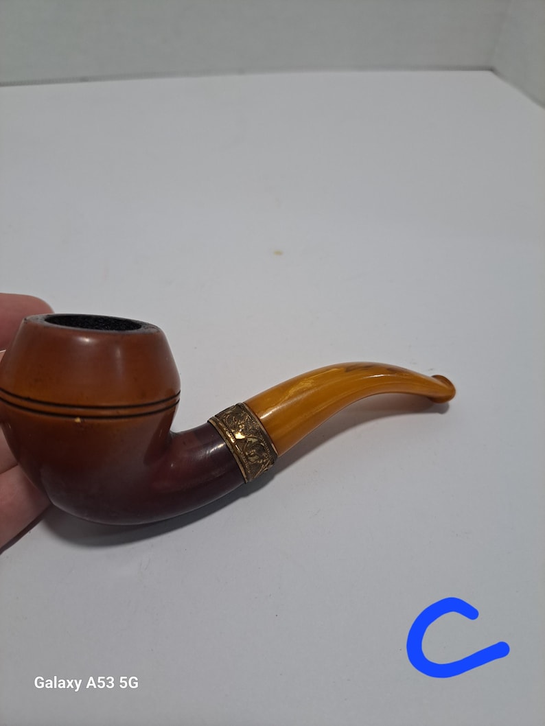 Op de afbeelding: Een bruine en amberkleurige pijp met een gouden band. De pijp heeft een gebogen steel en een kom met een bruin en amberkleurig patroon.