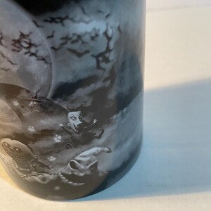 May include: A black and white ceramic mug featuring a graphic print of characters from The Nightmare Before Christmas, including a large moon, flying bats, and figures in a dark, cloudy, and atmospheric night scene with a detailed illustration.