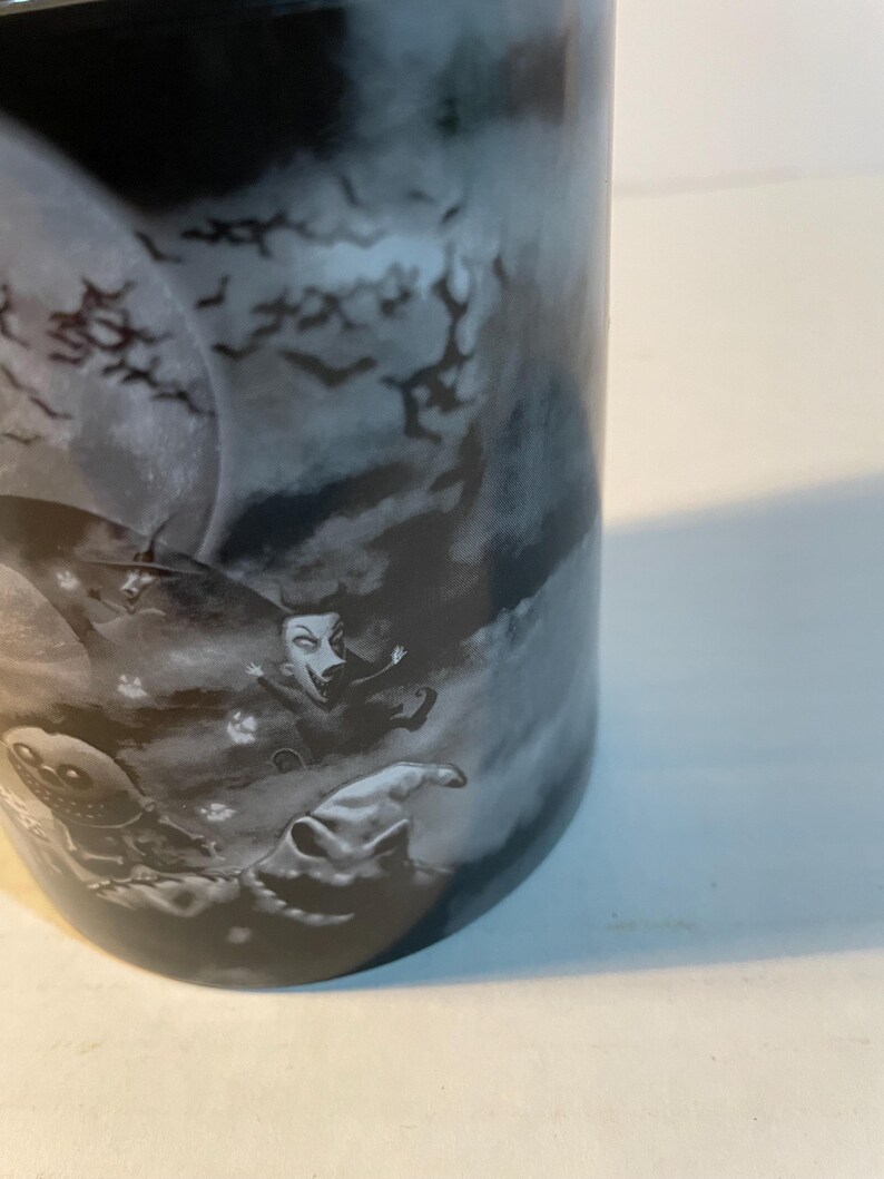 May include: A black and white ceramic mug featuring a graphic print of characters from The Nightmare Before Christmas, including a large moon, flying bats, and figures in a dark, cloudy, and atmospheric night scene with a detailed illustration.