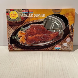 May include: A rectangular product box for a Nordic Ware Sizzler Server. The box features an image of a cooked steak on an oval metal platter with a black holder. Text on the box includes 'SERVO KING SIZZLER SERVER' and 'Durable Bakelite Holder'.